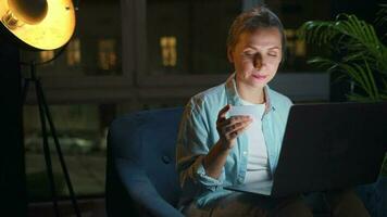 Woman is sitting in the armchair and makes an online purchase using a credit card and laptop at night video