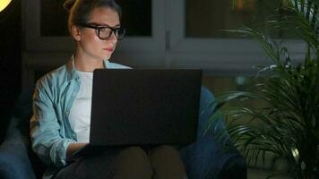 mulher com óculos é sentado dentro a poltrona e trabalhando em uma computador portátil às noite. conceito do controlo remoto trabalhos video