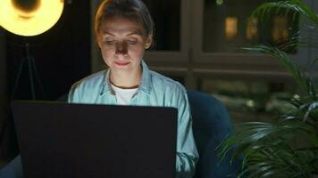 mulher é sentado dentro a poltrona e trabalhando em uma computador portátil às noite. conceito do controlo remoto trabalhos video
