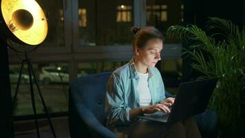 Woman is sitting in the armchair and working on a laptop at night. Concept of remote work video