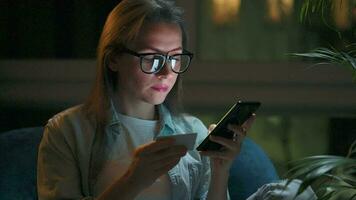 Woman is sitting in the armchair and makes an online purchase using a credit card and smartphone at night video