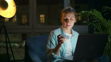 Woman is sitting in the armchair and makes an online purchase using a credit card and laptop at night video