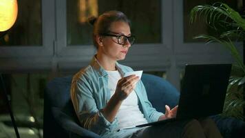 Woman is sitting in the armchair and makes an online purchase using a credit card and laptop at night video