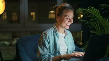 sonriente mujer es sentado en el Sillón y trabajando en un ordenador portátil o chateando con alguien a noche. concepto de remoto trabajo video