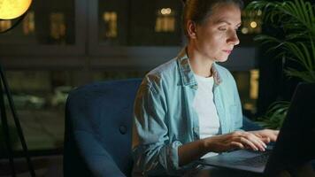 Woman is sitting in the armchair and working on a laptop at night. Concept of remote work video