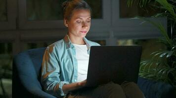 Woman is sitting in the armchair and makes an online purchase using a credit card and laptop at night video