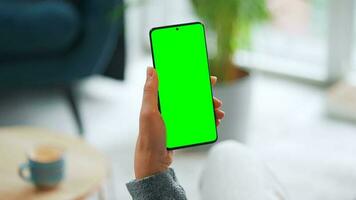 Woman at home using smartphone with green mock-up screen in vertical mode. Girl browsing Internet, watching content video