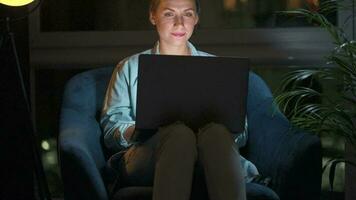 mujer es sentado en el Sillón y hace un en línea compra utilizando un crédito tarjeta y ordenador portátil a noche video