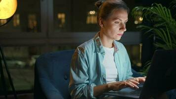 Woman is sitting in the armchair and working on a laptop at night. Concept of remote work video