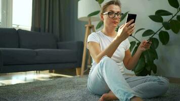 femme avec des lunettes est séance sur le tapis et fait du un en ligne achat en utilisant une crédit carte et téléphone intelligent. en ligne achats video