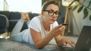 Woman with glasses is lies on the floor and makes an online purchase using a credit card and smartphone. Online shopping video