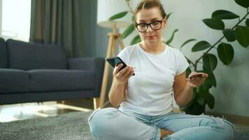 mujer con lentes es sentado en el alfombra y hace un en línea compra utilizando un crédito tarjeta y teléfono inteligente en línea compras video