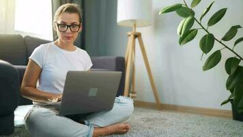 vrouw met bril is zittend Aan de tapijt en werken Aan een laptop. een pluizig kat leugens Aan de bankstel achter haar. concept van afgelegen werk video