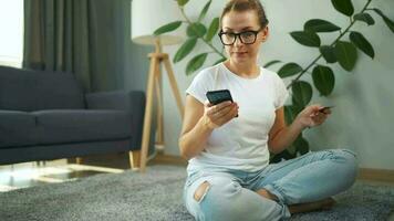 femme avec des lunettes est séance sur le tapis et fait du un en ligne achat en utilisant une crédit carte et téléphone intelligent. en ligne achats video