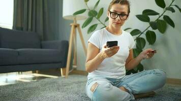 mujer con lentes es sentado en el alfombra y hace un en línea compra utilizando un crédito tarjeta y teléfono inteligente en línea compras video