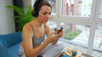 Lycklig avslappnad kvinna i trådlös hörlurar lyssnande till musik och använder sig av mobil appar eller kommunicerar på social nät på smartphone video