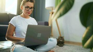 donna con bicchieri è seduta su il tappeto e Lavorando su un' il computer portatile. un' soffice gatto bugie su il divano dietro a suo. concetto di a distanza opera video