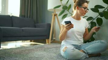 Woman with glasses is sitting on the carpet and makes an online purchase using a credit card and smartphone. Online shopping video