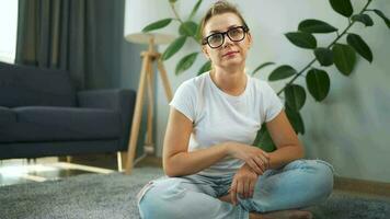 mulher com óculos olhando às a Câmera sentado em a tapete dentro a interior do uma acolhedor apartamento video