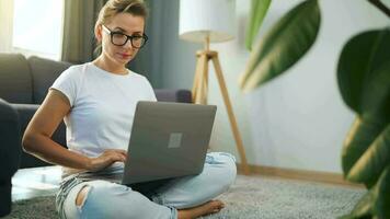 Frau mit Brille ist Sitzung auf das Teppich und Arbeiten auf ein Laptop. ein flauschige Katze Lügen auf das Couch hinter ihr. Konzept von Fernbedienung Arbeit video