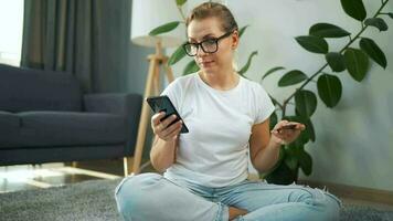 mujer con lentes es sentado en el alfombra y hace un en línea compra utilizando un crédito tarjeta y teléfono inteligente en línea compras video