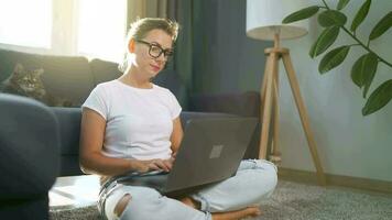 Frau mit Brille ist Sitzung auf das Teppich und Arbeiten auf ein Laptop. ein flauschige Katze Lügen auf das Couch hinter ihr. Konzept von Fernbedienung Arbeit video