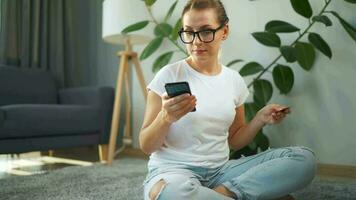 Woman with glasses is sitting on the carpet and makes an online purchase using a credit card and smartphone. Online shopping video