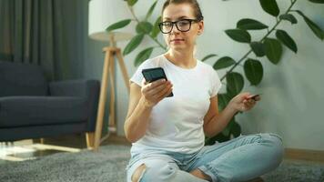 Woman with glasses is sitting on the carpet and makes an online purchase using a credit card and smartphone. Online shopping video