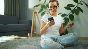 mujer con lentes es sentado en el alfombra y hace un en línea compra utilizando un crédito tarjeta y teléfono inteligente en línea compras video