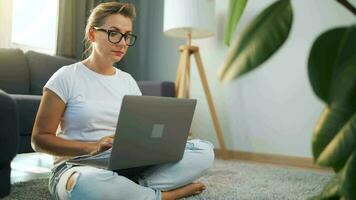 donna con bicchieri è seduta su il tappeto e Lavorando su un' il computer portatile. un' soffice gatto bugie su il divano dietro a suo. concetto di a distanza opera video