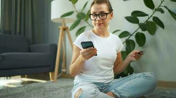 Woman with glasses is sitting on the carpet and makes an online purchase using a credit card and smartphone. Online shopping video