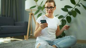 vrouw met bril is zittend Aan de tapijt en maakt een online aankoop gebruik makend van een credit kaart en smartphone. online boodschappen doen video