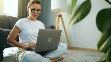 donna con bicchieri è seduta su il tappeto e Lavorando su un' il computer portatile. un' soffice gatto bugie su il divano dietro a suo. concetto di a distanza opera video