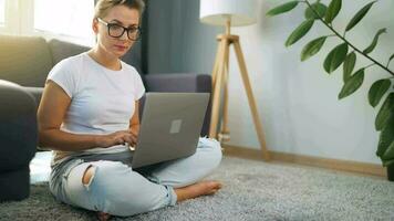 Frau mit Brille ist Sitzung auf das Teppich und Arbeiten auf ein Laptop. ein flauschige Katze Lügen auf das Couch hinter ihr. Konzept von Fernbedienung Arbeit video