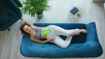 Overhead shot of relaxed woman in wireless headphones listening to music and using mobile apps on smartphone with green mock-up screen video