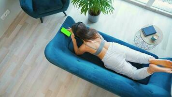 Overhead shot of relaxed woman in wireless headphones listening to music and using mobile apps on smartphone with green mock-up screen video