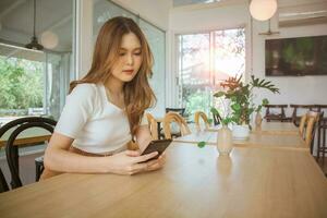 mujer asiática feliz adulta joven que usa un teléfono inteligente para la aplicación de medios sociales, compra en línea, transfiere dinero, finanzas, mensajes en la cafetería. fondo con ventana y luz solar cálida en la temporada de verano. foto