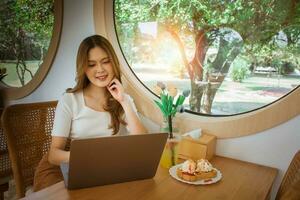 joven inteligente asiático mujer Lanza libre en línea trabajando desde café con ordenador portátil computadora. joven hermosa alegre hembra sonriente mientras laboral, pensando acerca de cómo a tomar el negocio a tecnológico alturas foto