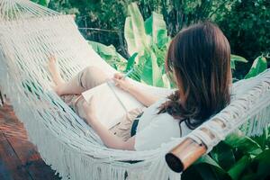 Beautiful Asian woman wearing glasses taking note on white hammock in garden at home. view from behind. morning sun. Slow living, weekend leisure activity. Quarantine and self isolation period. photo