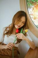 retrato de bonito dama participación tulipanes en manos. joven romántico mujer cerca ojos sonriente y olfateando hermosa de flores, sentado satisfecho en cafetería. concepto de hembra belleza, primavera y feminidad. foto