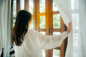 Side view of young woman wearing glasses wake up in bedroom at home or hotel, open white curtains look in window admiring to get fresh air. welcome new day morning feel positive and optimistic. photo