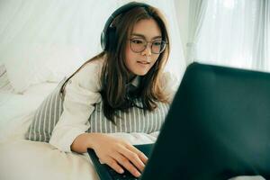 hermosa asiático joven mujer vistiendo lentes y auriculares escuchando música o acecho película con ordenador portátil en el blanco cama a hogar. bonito niña acostado abajo relajante y sonriente con cuaderno computadora. foto