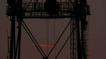 The plane flies leaving a contrail, it is bright orange due to the sunset. Silhouette of a metal structure in the foreground. Plane jet at cruising altitude, twilight glow video