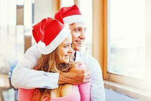 a couple wearing christmas santa red hats hugging and looking to the window. Focus on a woman photo