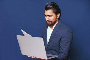 Young stylish Indian man in formals using laptop computer and reading from the document, over studio blue background photo