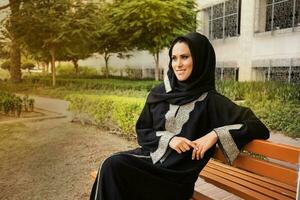 young Caucasian girl wearing hijab and jilbab sitting on a bench smiling in a park photo