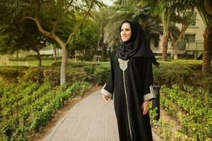 joven caucásico niña vistiendo hijab y jilbab en pie y sonriente en un parque foto