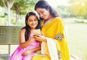 indio madre y hija en tradicional étnico ropa utilizando móvil teléfono en un banco de el parque en Delhi foto