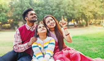 Beautiful Indian family sitting on a grass in the park, pointing at something and looking up photo