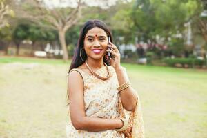 hermosa indio mujer en sari hablando en móvil teléfono, en pie en el parque foto